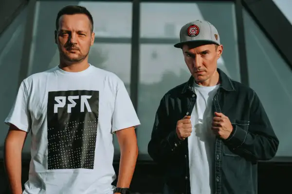 Dwóch członków Paktofoniki, jeden w białej koszulce z logo, drugi w czarnej kurtce i czapce z daszkiem.